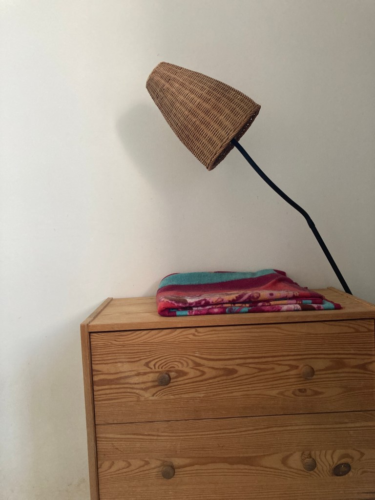 Une petite commode avec à droite un luminaire. la tête de la lampe est très penchée au-dessus de la commode comme si on l’avait tordue dans cet angle.