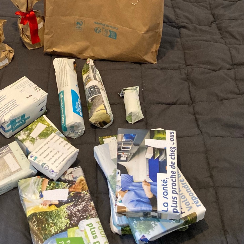 Sur une couverture noire, divers cadeaux emballés dans des feuilles de catalogue. Deux cadeaux cylindriques sont emballés par une grande feuille de papier maintenue par un ruban. Il y a également un grand sac de papier fermé à l’aide de ficelle. Chaque paquet cadeau a une étiquette nominative en braille.