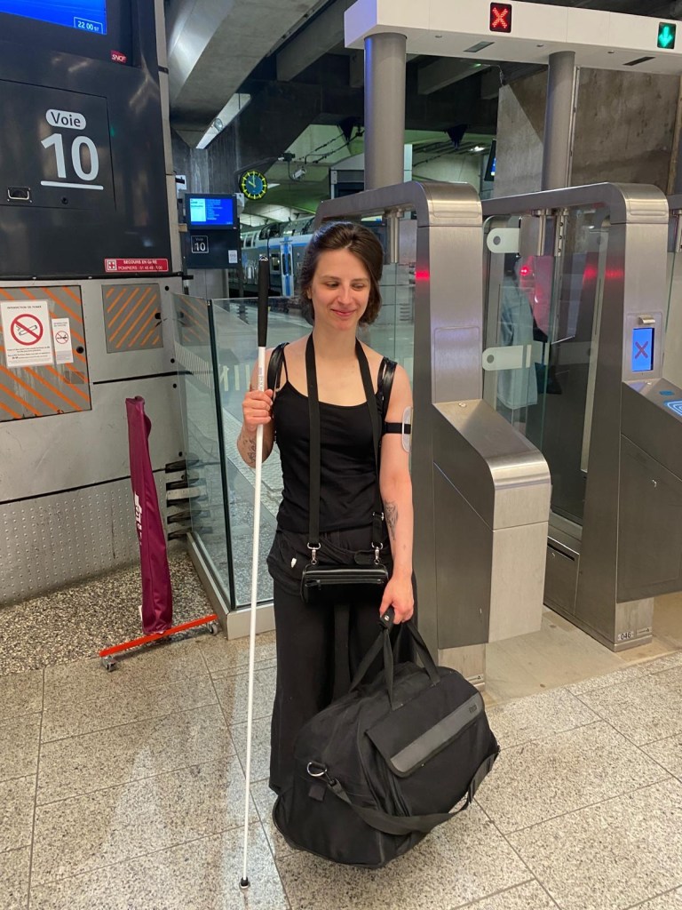 Bibi est dans le hall de la gare M*ntparnasse. Elle porte un sarouel noir et une petite natte. Elle sourit. Dans sa main gauche, un gros sac de voyage noir. Dans sa main droije, une très grande canne blanche qui lui arrive au niveau du front.