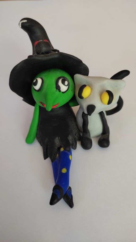 Une figurine en pâte Fimo représentant une sorcière et un chat. La peau de la sorcière est verte. Elle porte une robe noire dont le bas est élimé, un chapeau pointu noir sur lequel est posé une araignée blanche, des collants bleus à pois jaunes et des souliers noirs à boucle. Elle a un nez crochu, pas de bouche et de grands yeux bleus qui fixent la personne qui l’observe. Son bras droit enlace un chat à sa droite. Le chat a les yeux jaunes, son pelage est gris hormis les oreilles, les pattes et la queue qui sont noires.