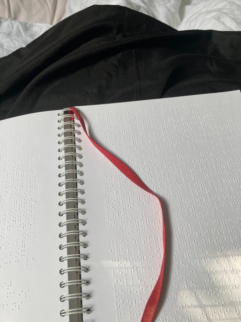 Sur un fond noir: le livre en braille L’Institut de Stephen King est ouvert. Les pages sont au format A4 et la reliure est en spirale. Un cordon rouge est relié aux spirales et posé sur la page de droite.