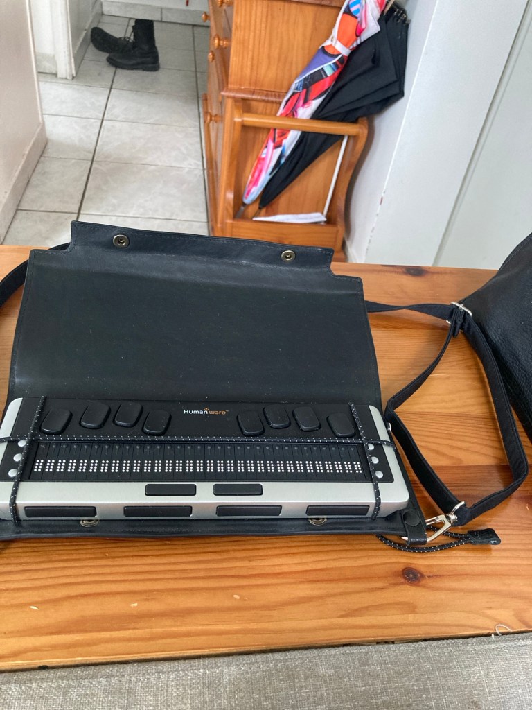 Une HumanWare Briailliant BI de 32 cellules. Il y a un clavier dit Perkins tout en haut, des boutons de chaque côté de l’afficheur brille tout en bas. Elle est maintenue dans sa sacoche par trois élastiques.