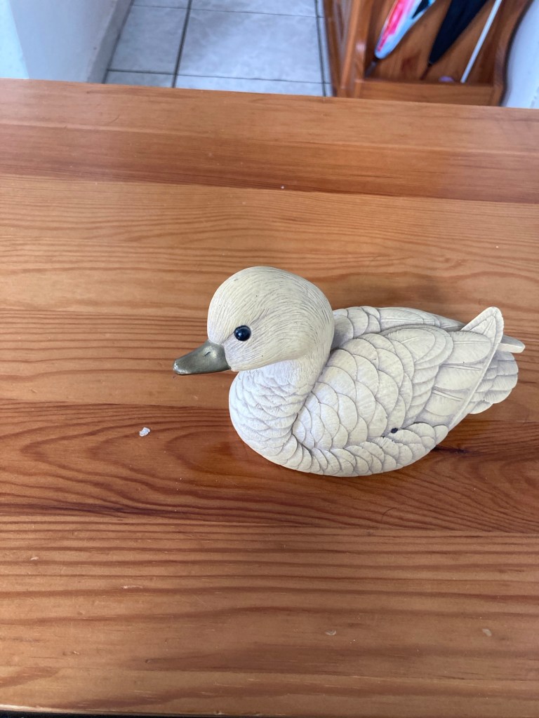 La figurine d’un canard sculpté dans du bois clair. Les plumes sont détaillées et on les sent au toucher. Le bec est en métal.