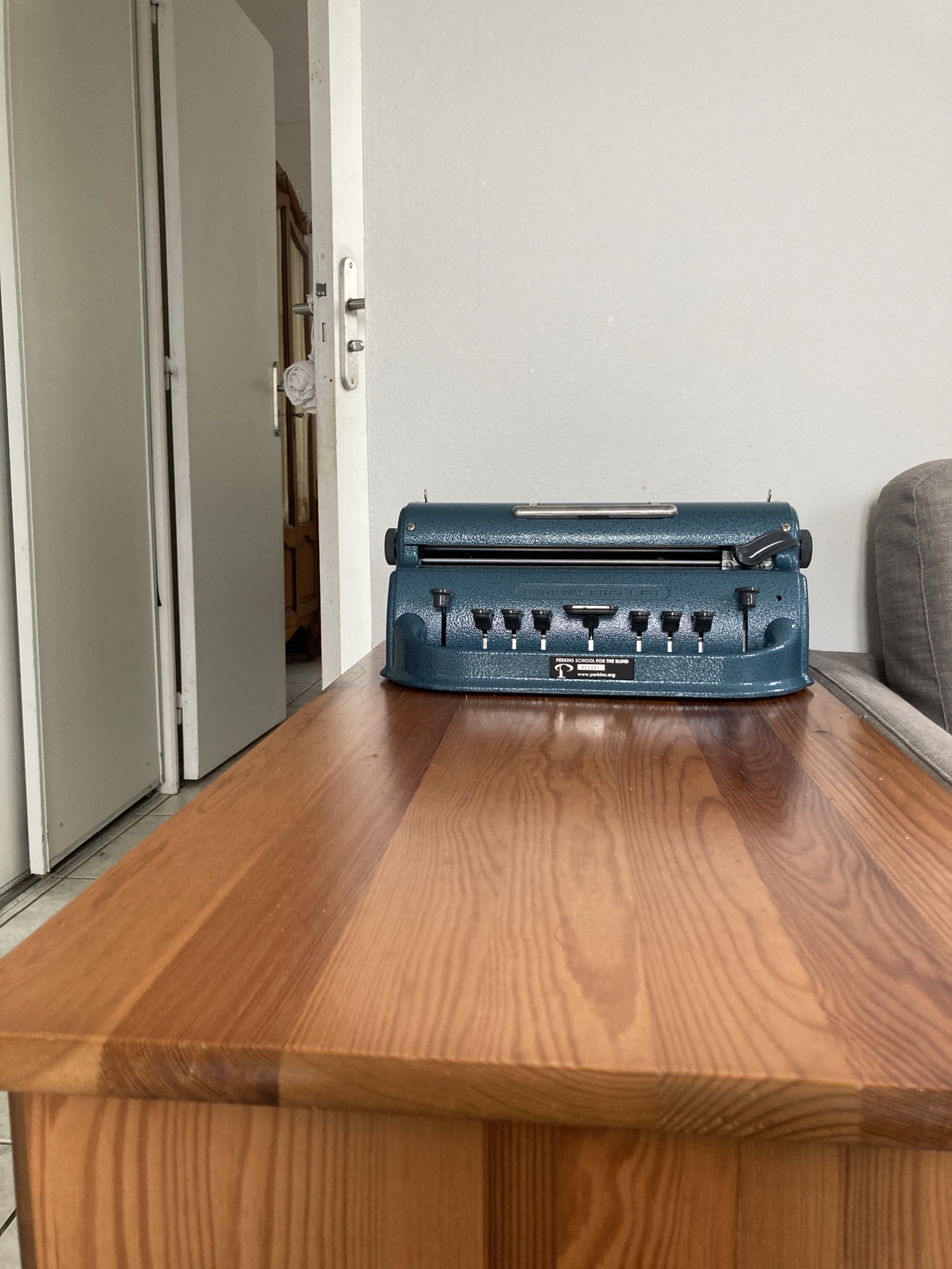 Sur une commode en bois clair est posée une machine à écrire le braille Perkins de couleur bleue.
