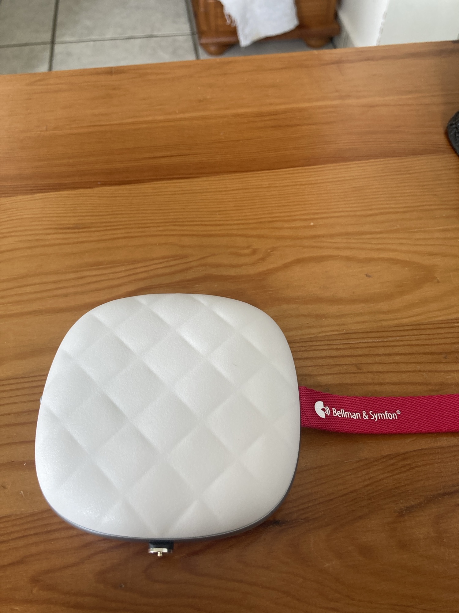 Un carré de texture matelassée blanc. Sur sa tranche gauche il y a un interupteur. Sur celle du milieu il y a un embout de charge magnétique. Sur celle de droite il y a une sangle.