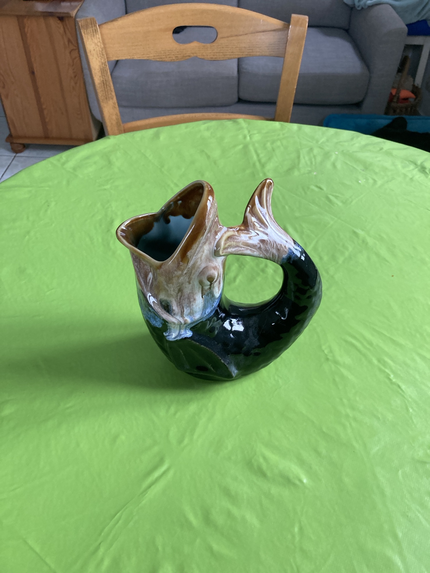 Une cruche en céramique en forme de poisson est positionnée au centre sur une table ronde recouverte d’une nappe verte. La cruche a une finition brillante, avec un dégradé de couleurs passant du brun et du beige en haut au noir à la base. La bouche du poisson est ouverte, formant le bec, et sa queue se courbe vers le haut pour former une poignée.