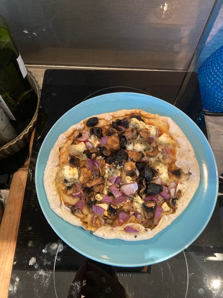 Sur une assiette ronde bleue est posée une pizza maison aux champigons, fromage de chèvre, olives noires et oignon.