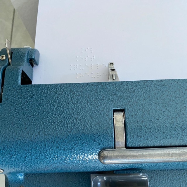 Gros plan sur une feuille de papier blanche insérée dans une machine à écrire Perkins bleue. Sur la feuille est écrit en braille: « Il Est Quelle Heure ».