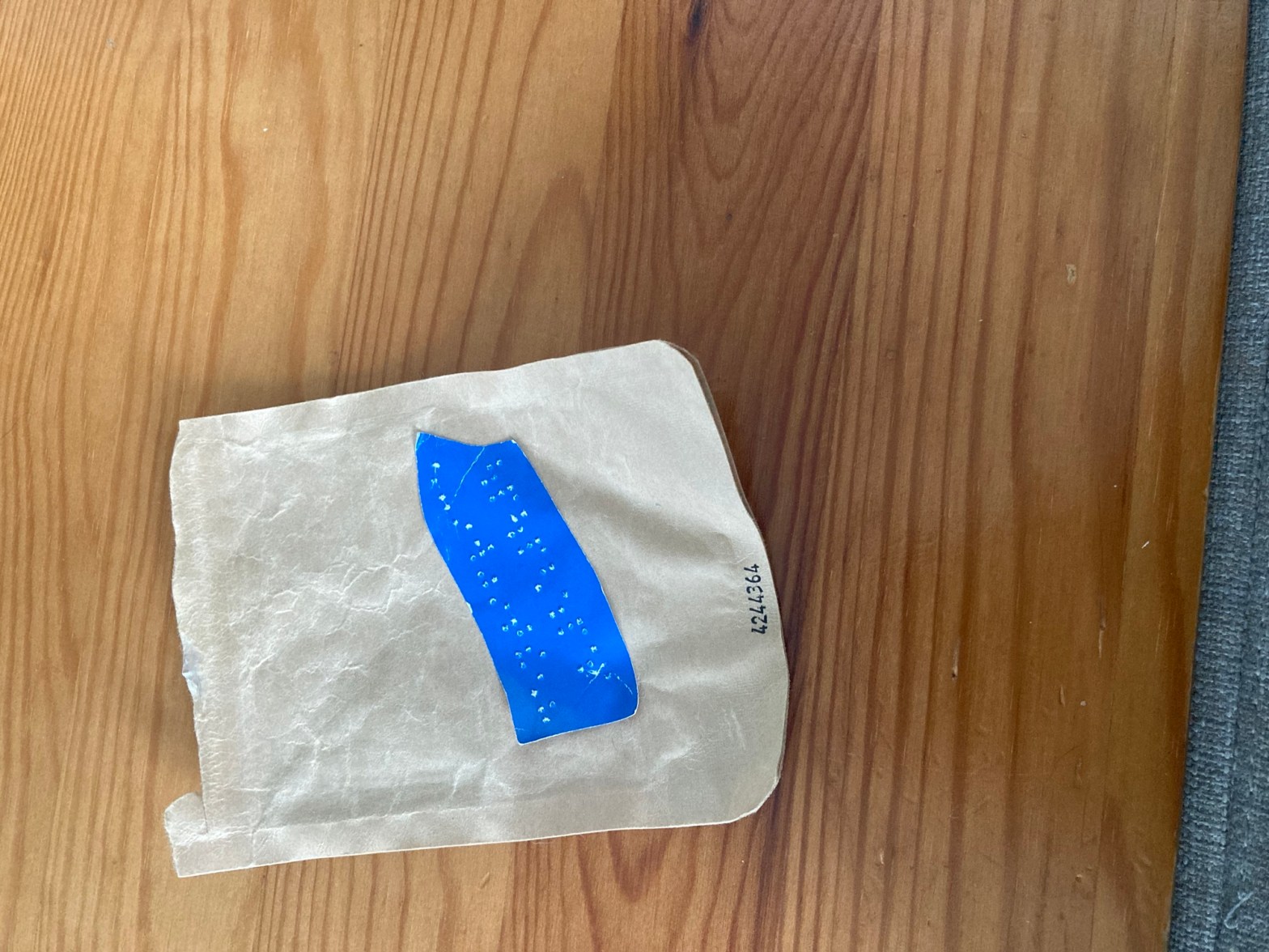 Sur un meuble en bois est posé un sachet de dentifrice en pastilles vide. Sur le sachet, une étiquette braille (réutilisation du papier d’un prospectus) a été collée.