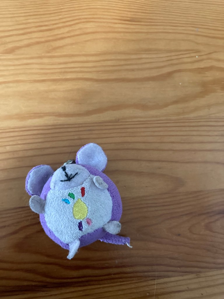 Petite peluche à l’effigie d’une souris violette souriante. La souris est en forme de boule. Sur son ventre est brodé le logo du Téléthon.