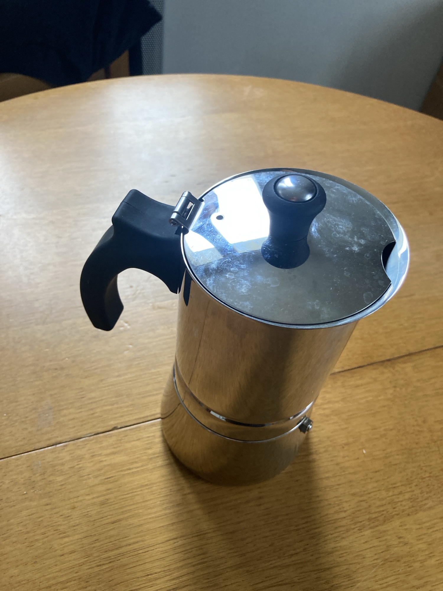 Une cafetière italienne est posée sur une table en bois. Elle est cylindrique, en métal, avec une poignée noire sur sa gauche et un bouton noir sur le couvercle.