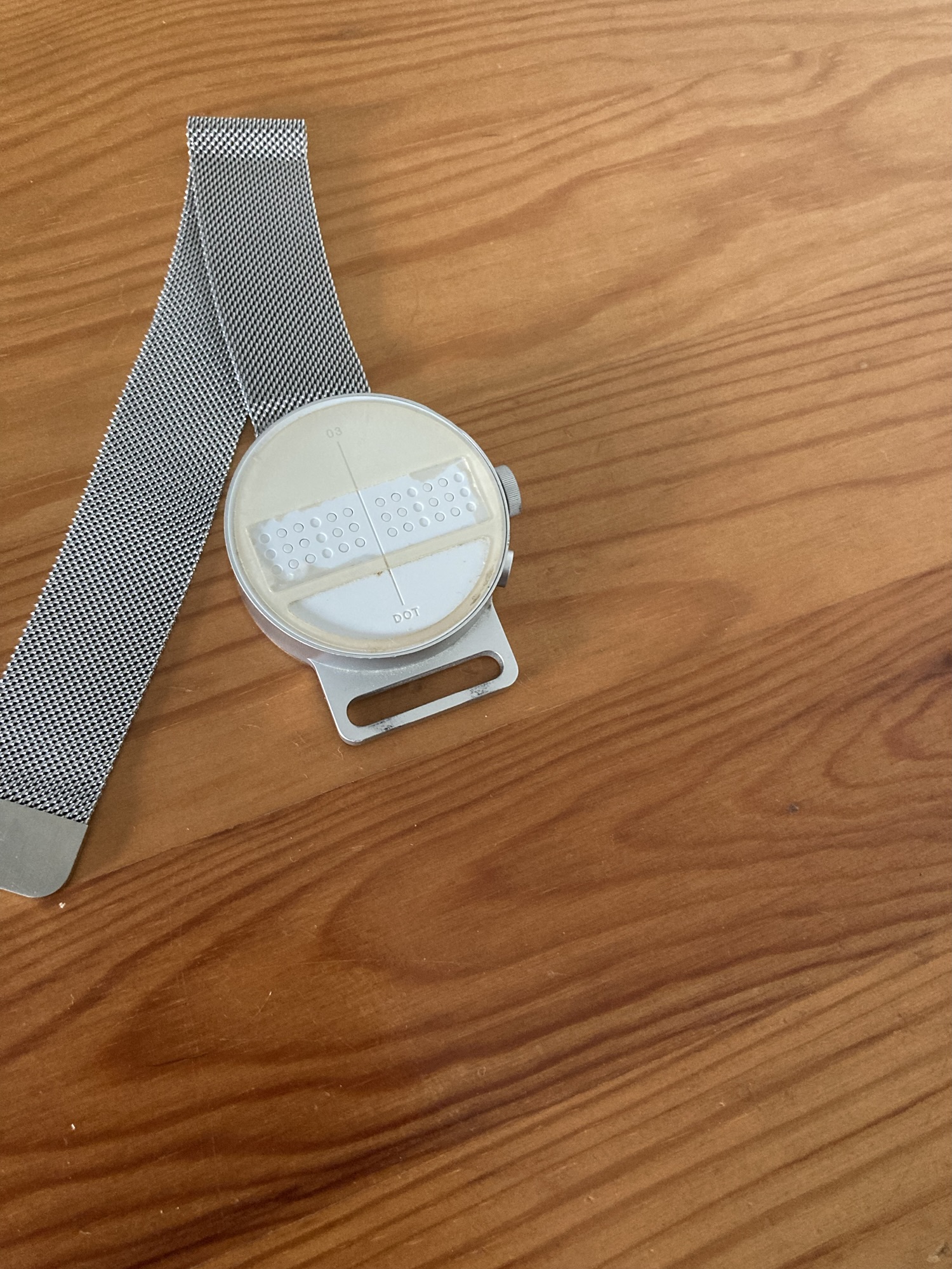 Une montre avec une sangle en maille posée sur une surface en bois. La montre possède un cadran blanc et rond avec un design unique comportant une grille de points de braille ainsi qu'une zone lisse en-dessous. Le bracelet est métallique et semble flexible, s’étendant du côté gauche du cadran. Le cadran de la montre a trois boutons sur le côté droit.